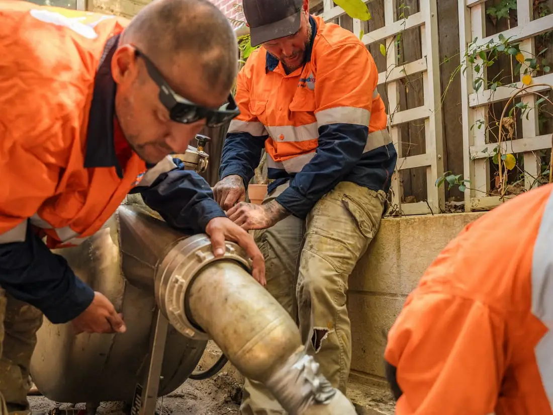 Pipe relining under slab in Perth starts outside the house: three of the crew guiding a liner from the inversion drum into the sewer pipe through a hand-dug access hole in a side passage.